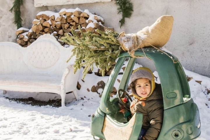 Child in a toy car with a Christmas tree on snowy ground, vintage couch and sign in background.