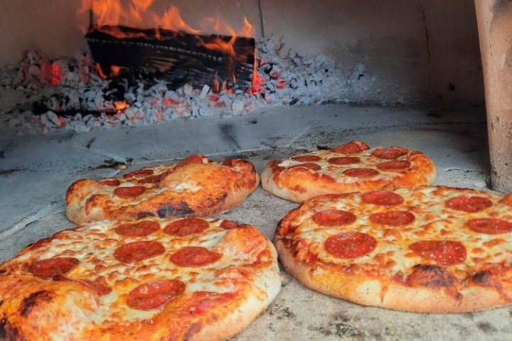 Three pepperoni pizzas baking in a wood-fired oven with flames.