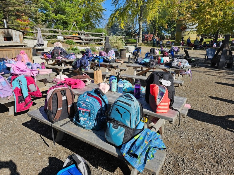Picnic tables with backpacks, jackets, and water bottles in a sunny outdoor park area.