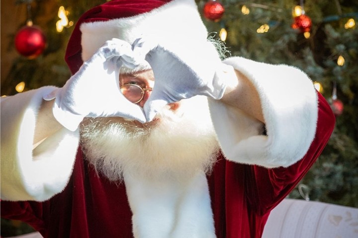 Santa making heart shape with hands, in front of decorated Christmas tree.