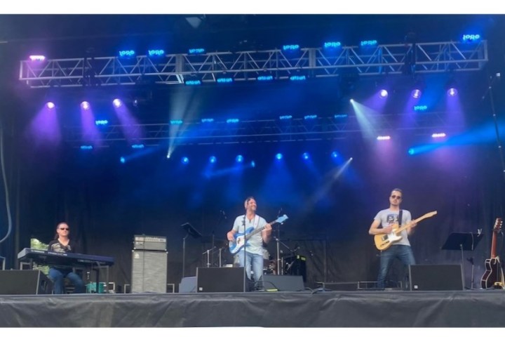Three musicians on stage with colorful lights, playing keyboard, bass guitar, and electric guitar.