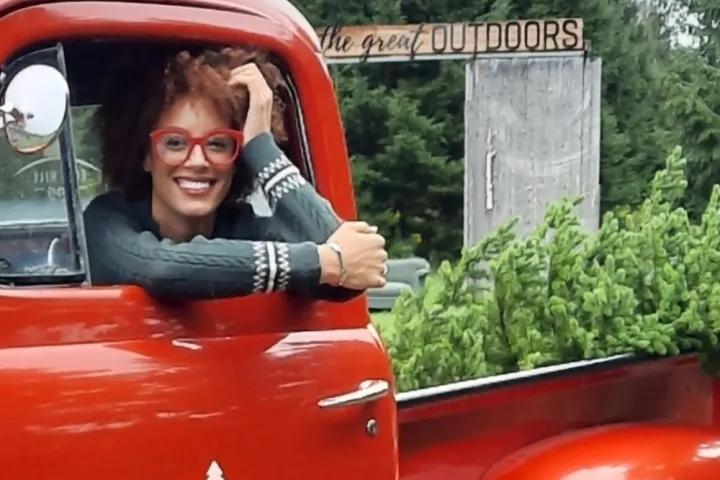 Person with glasses in red truck smiling, holding a Christmas tree, surrounded by pine trees.
