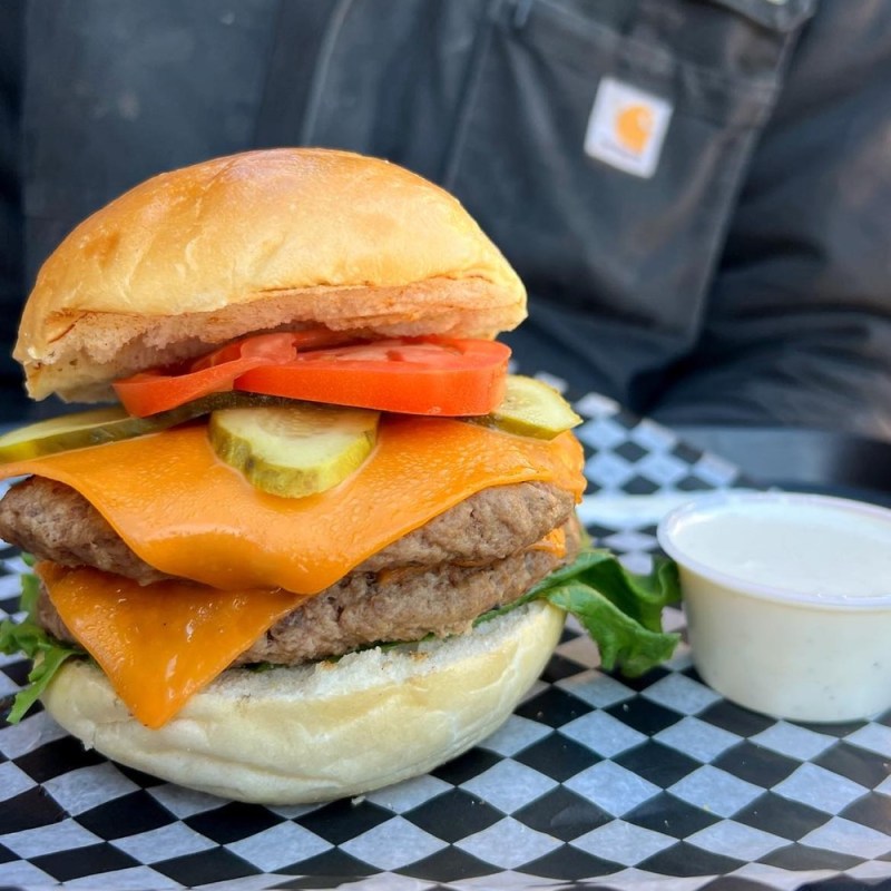 Cheeseburger with lettuce, tomato, and pickles on a checkered paper, with a sauce cup beside it.