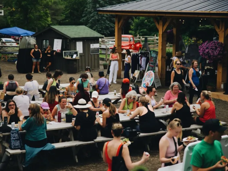 Crowd of people at outdoor picnic tables, enjoying food under a pavilion.