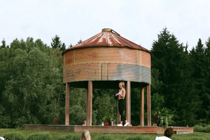 Outdoor yoga class with instructor on a stage and participants on mats on the grass.