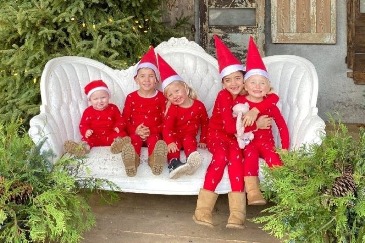 Five children in red pajamas and elf hats sitting on a white couch with Christmas trees nearby.