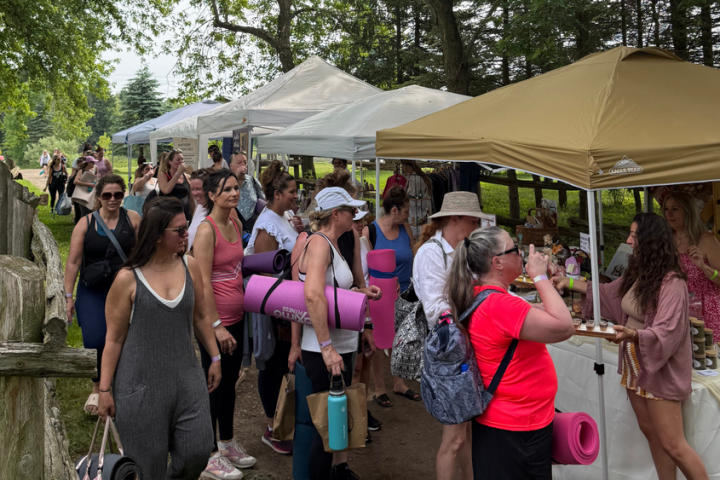People at outdoor market with yoga mats, browsing booths under white and tan tents on a sunny day.