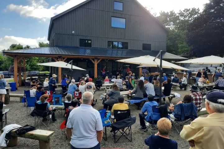 Outdoor gathering with people seated, facing a stage near a large building.