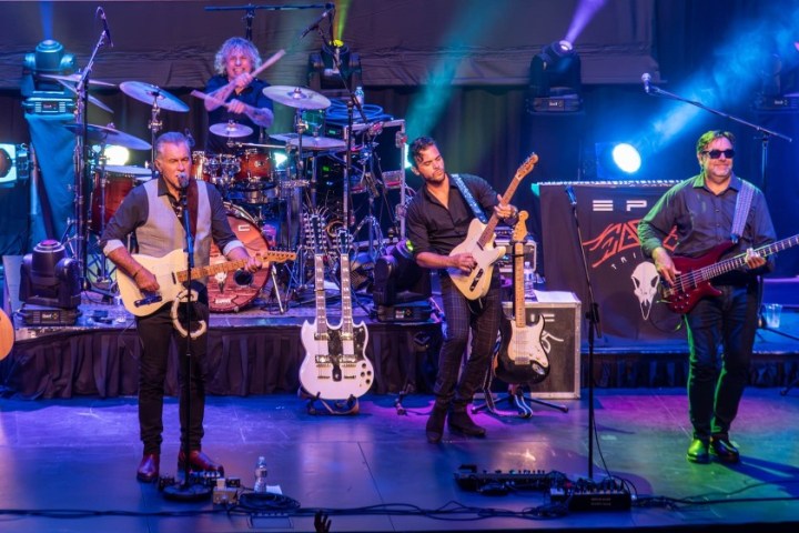 A band of four musicians performing on stage with guitars and drums under colorful lights.