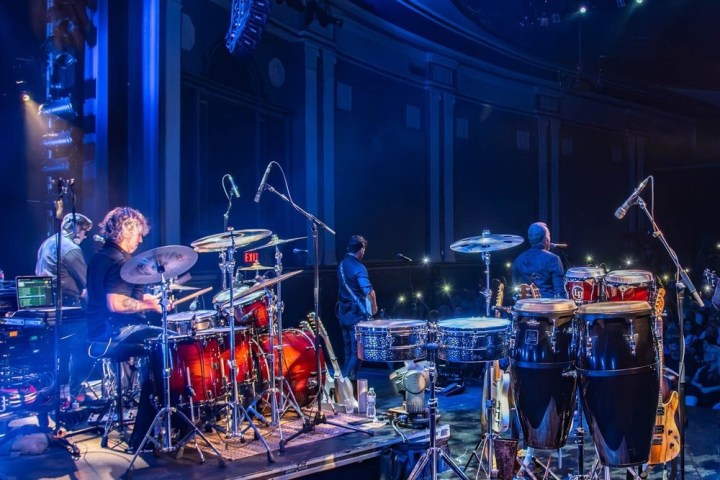 Band performing live on stage with drum kit and percussion instruments visible.