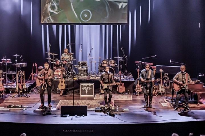 Band performing on stage with guitars and drums, audience watching.