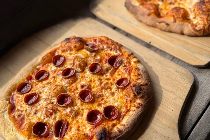 Two pepperoni pizzas on wooden paddles on a table.