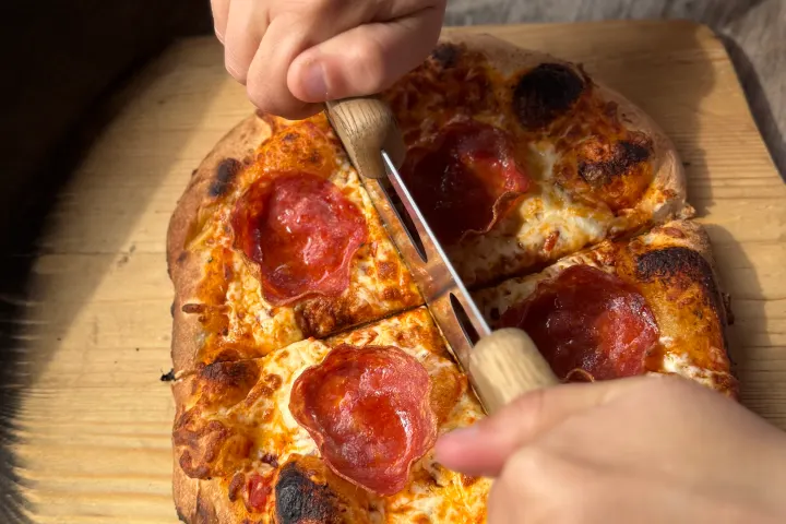 Hands cutting a pepperoni pizza on a wooden board with a double-handled knife.