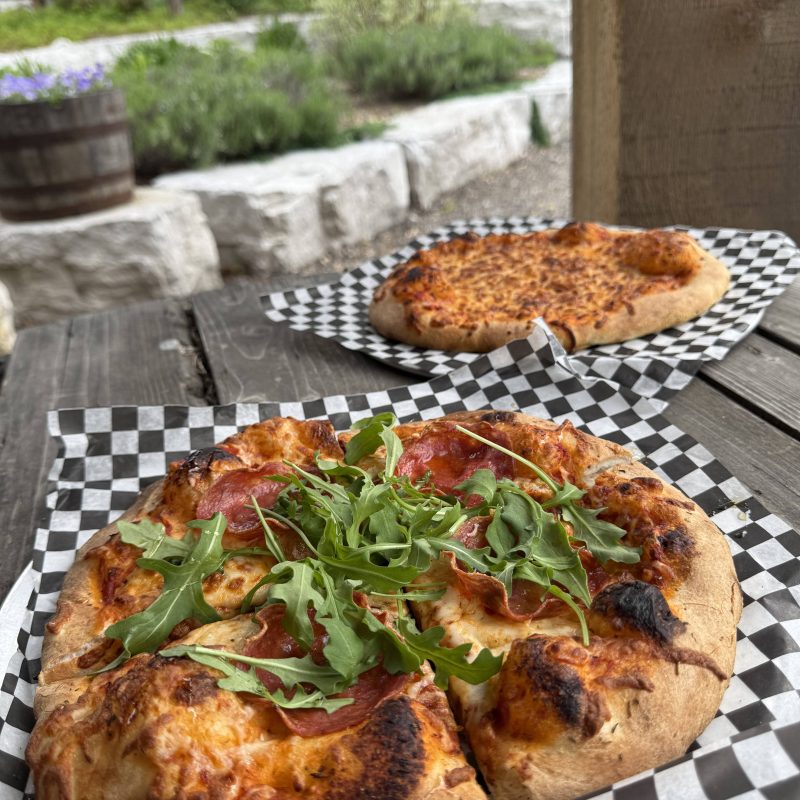Two pizzas with arugula and pepperoni on checkered paper outside on a wooden table.
