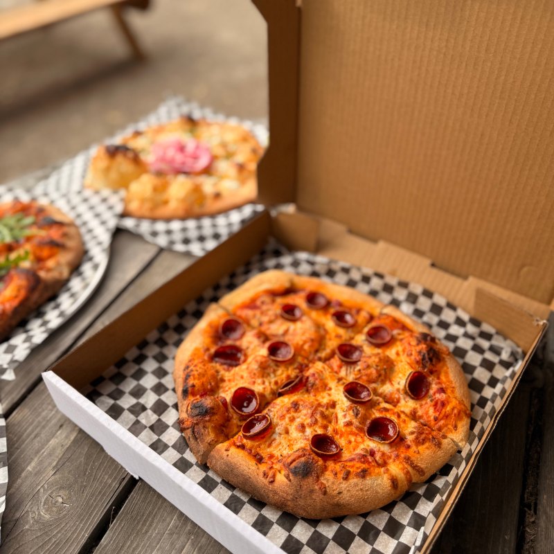 Pepperoni pizza in an open box on a picnic table, with other pizzas blurred in the background.