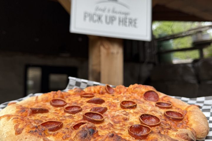 Pepperoni pizza on checkered paper with a 'Pick Up Here' sign in the background.