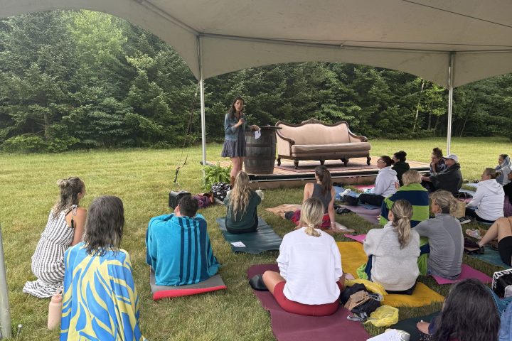 Outdoor gathering under tent with speaker and seated audience on grass.