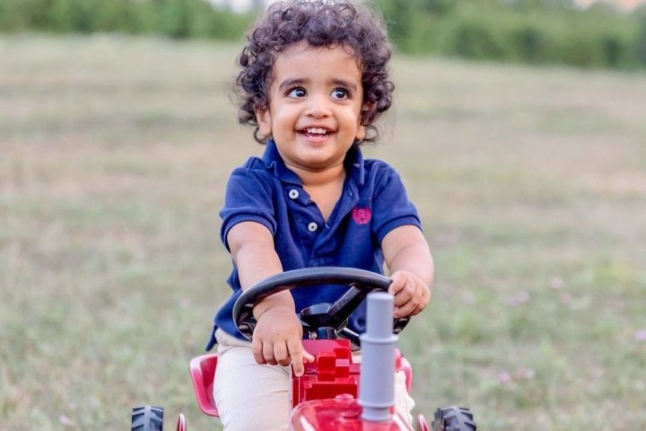 Smiling child in a navy shirt sits on a red toy tractor in a grassy field.