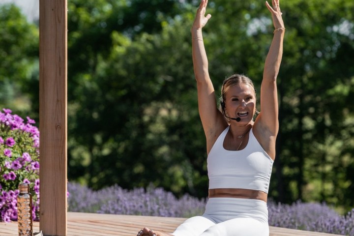 Person in white workout attire doing yoga outside with arms raised, surrounded by greenery and purple flowers.
