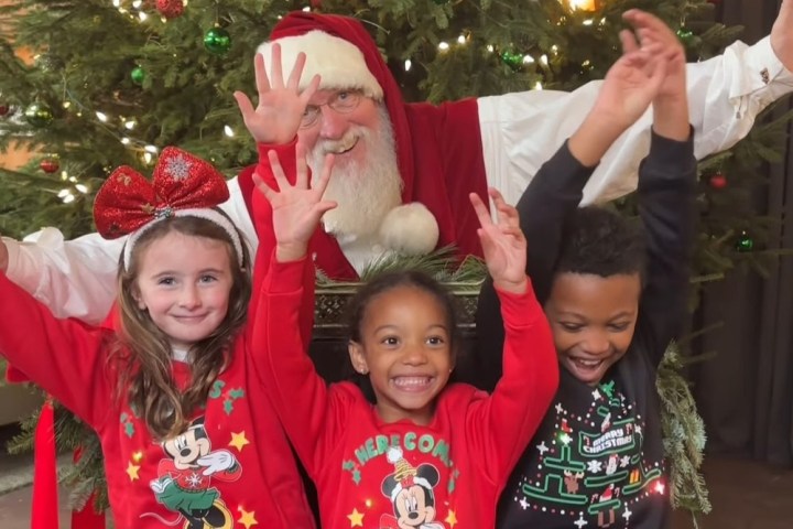Santa Claus with three smiling children raising hands in front of a Christmas tree.
