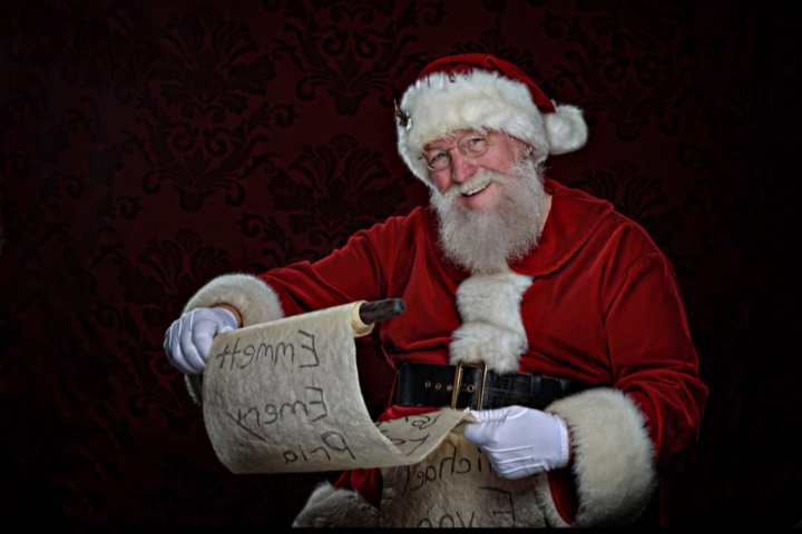 Santa Claus smiling, holding a long list, wearing traditional red suit with white trimming.