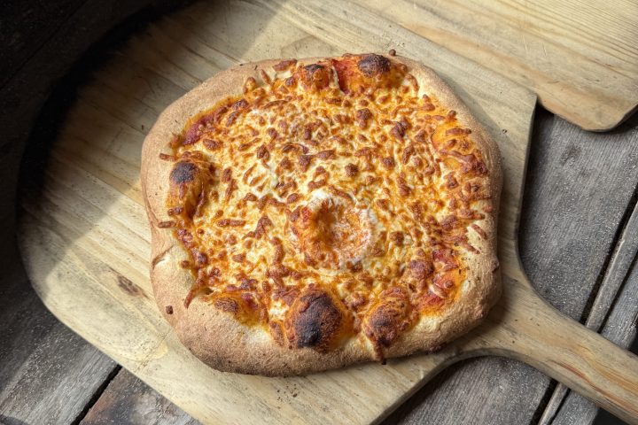 Cheesy pizza on a wooden paddle next to an oven.
