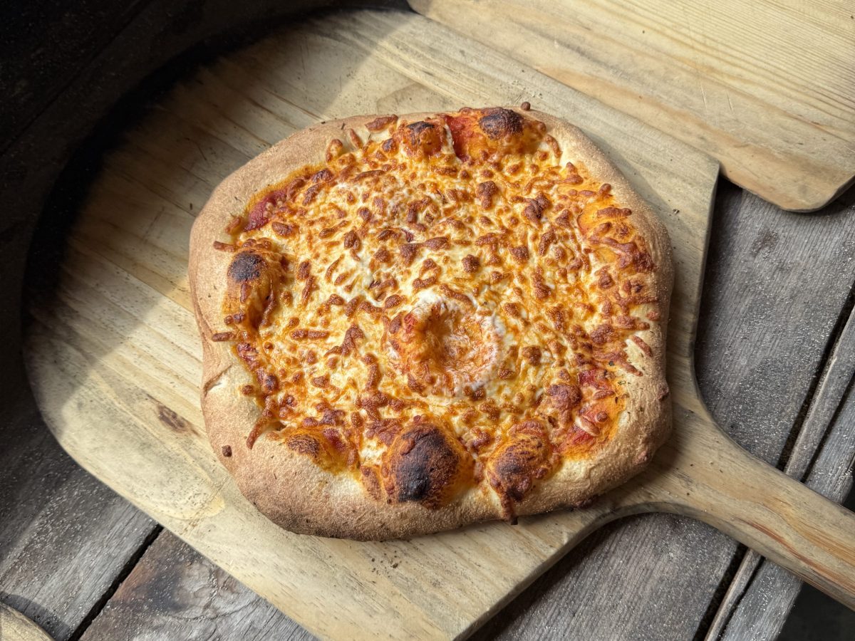 Cheesy pizza on a wooden paddle next to an oven.