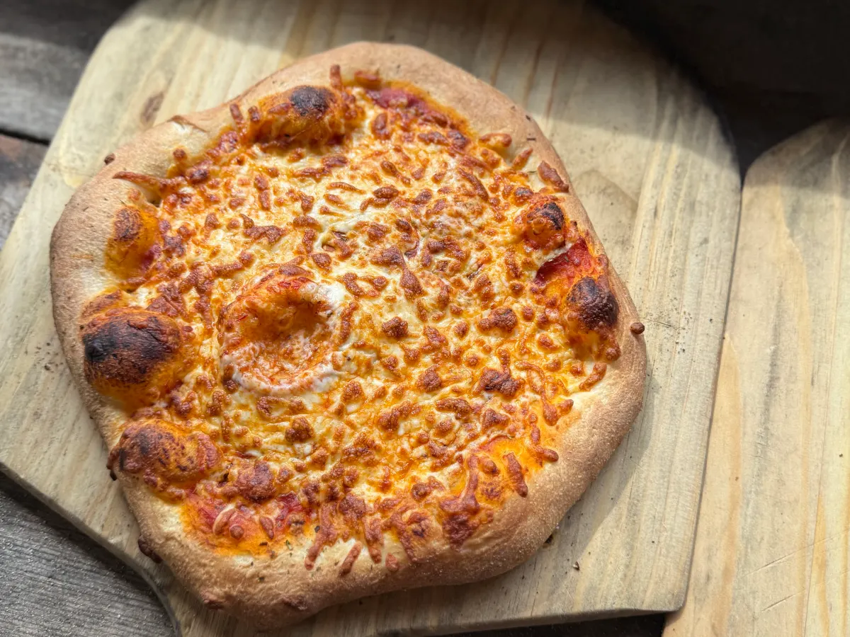 Cheesy pizza on a wooden paddle on a rustic wooden surface.