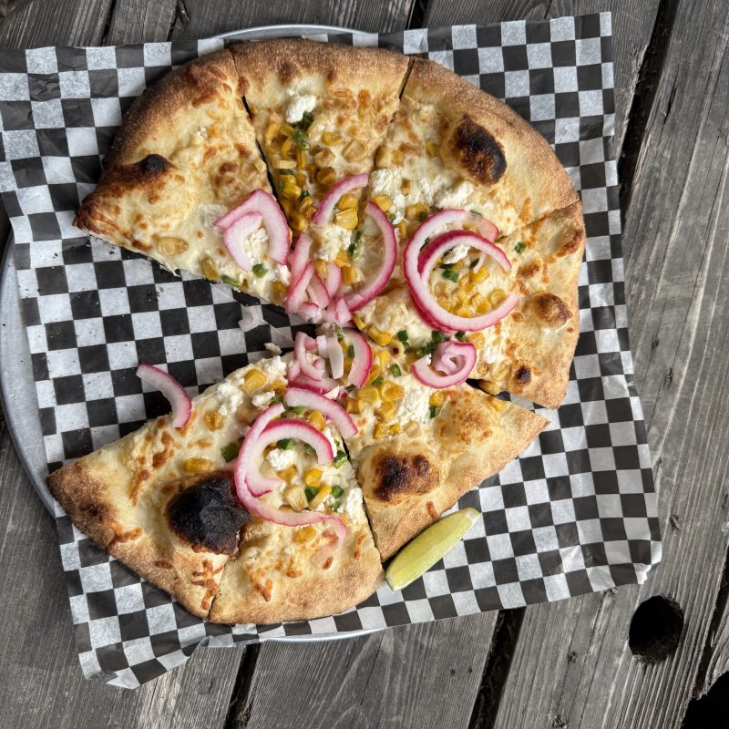 Craft pizza with pickled onions and corn on a checkered paper, on a wooden table.