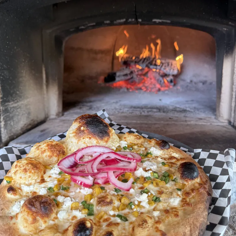 Wood-fired pizza with corn, onions, and cheese in front of a burning oven.