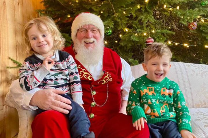 Two kids sit with Santa Claus on a couch in front of a decorated Christmas tree.