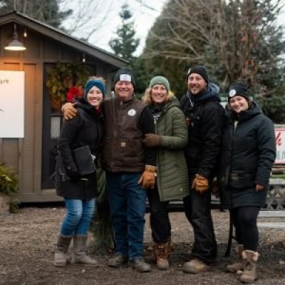 Group of five people in winter clothing smiling outside a rustic building with Christmas tree signs.
