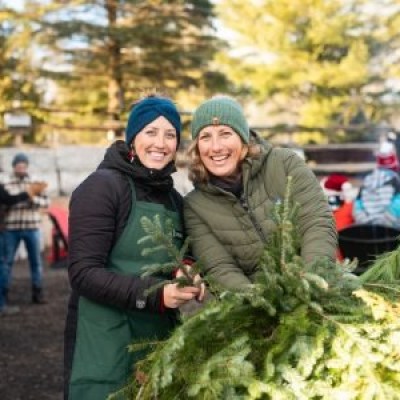 Two women smiling, holding fir branches outdoors in a winter setting.