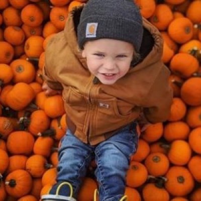 Child in jacket and beanie sitting on a pile of small pumpkins, smiling.