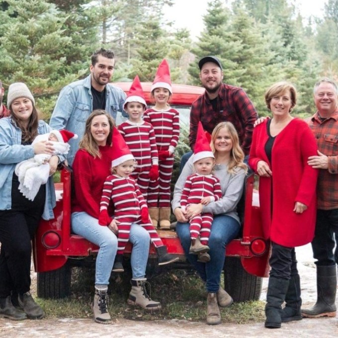 Family in winter clothing poses on a red truck surrounded by pine trees.