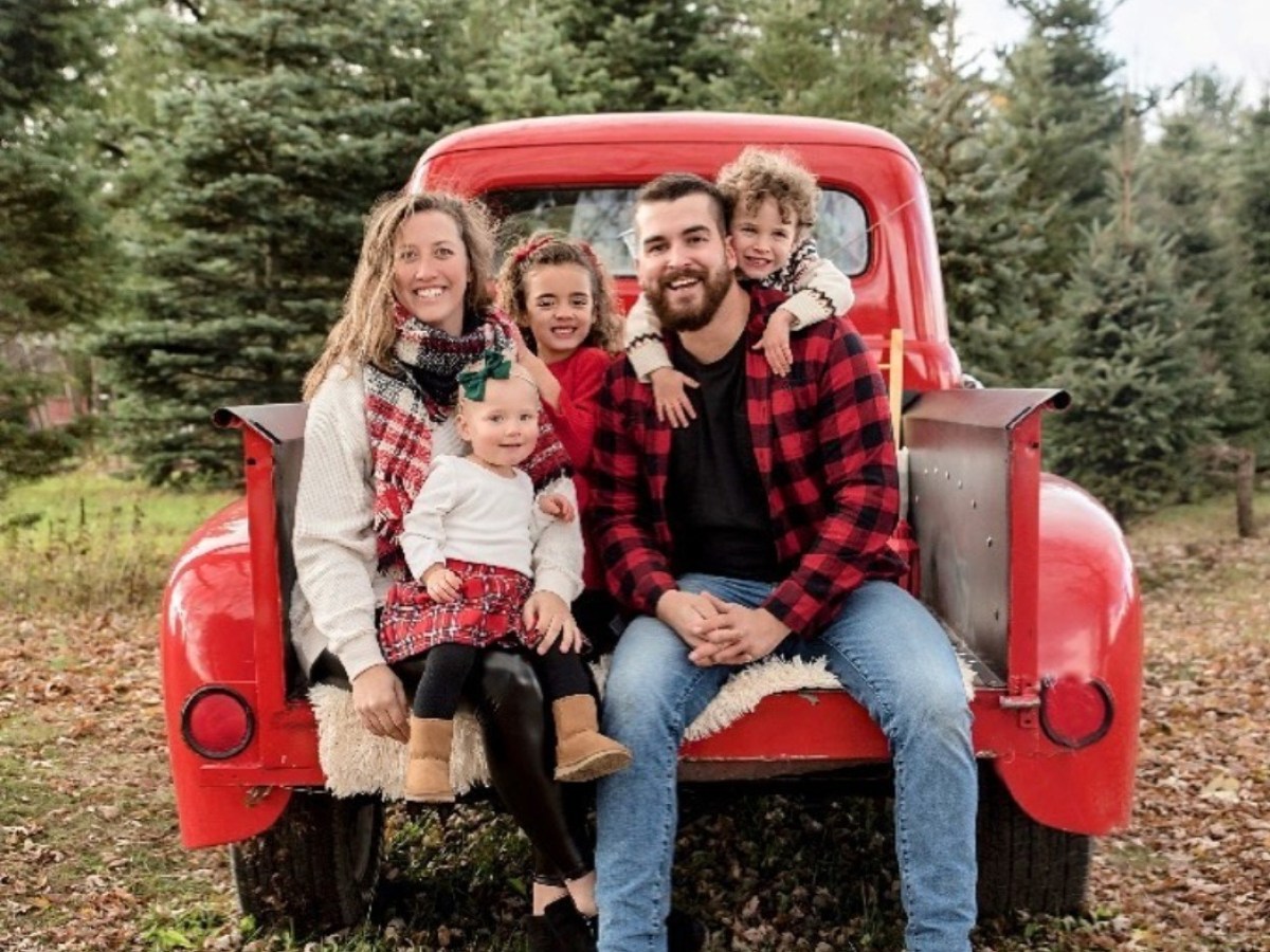 Family sitting on a red vintage truck with evergreen trees in the background.