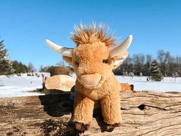 Plush toy cow with horns on wooden fence in snowy landscape under clear blue sky.
