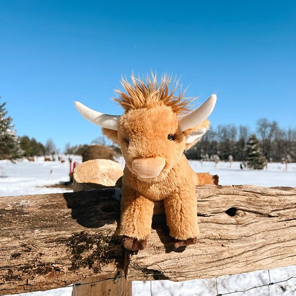 Plush toy cow with horns on wooden fence in snowy landscape under clear blue sky.