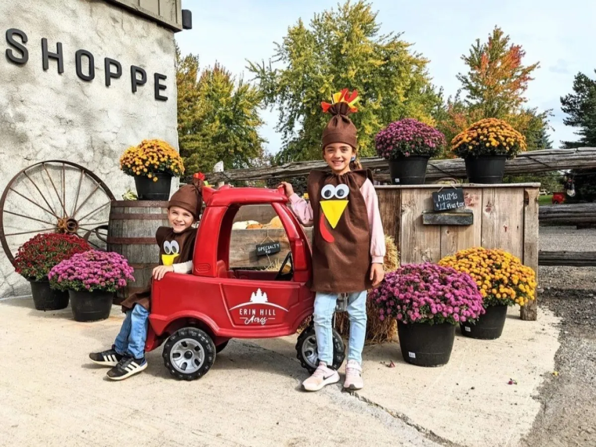 Two kids in turkey costumes by red toy car, surrounded by colorful potted flowers.