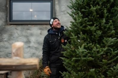Man in winter clothing smiles next to a large green Christmas tree outside a building with a window.