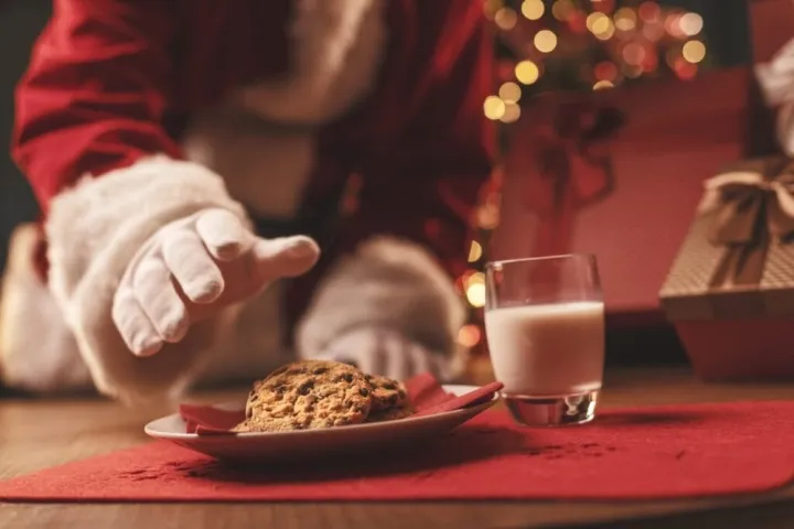 Santa reaching for a cookie on a plate with milk nearby, gifts in background.