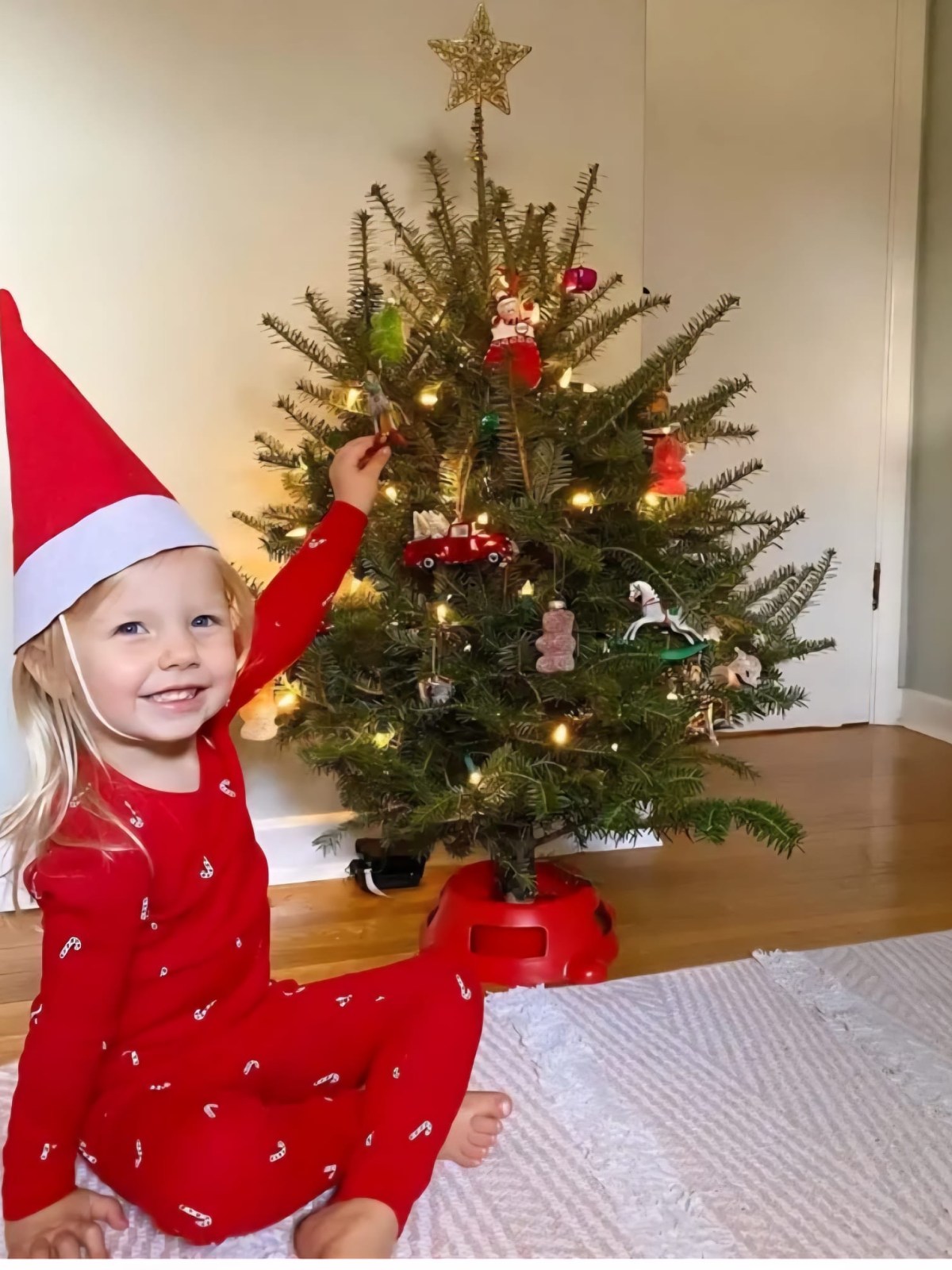 Child in red pajamas with Santa hat decorating small Christmas tree indoors.