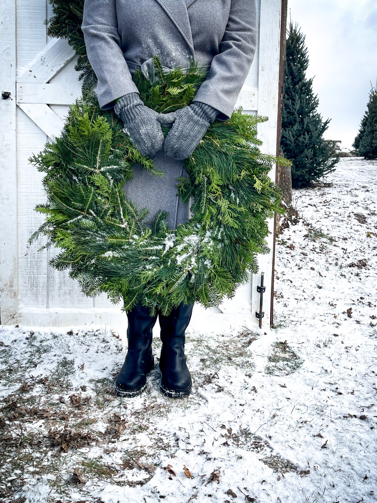 Person in gray coat holding green wreath, standing on snowy ground.