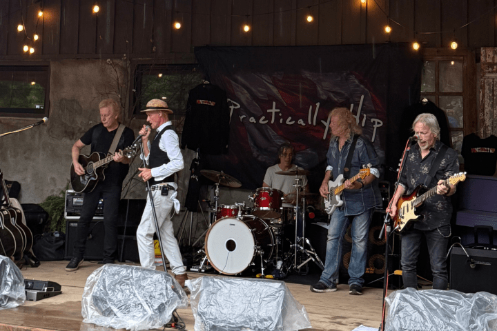 Band performing on outdoor stage with guitars, drums, and microphone. String lights are above.
