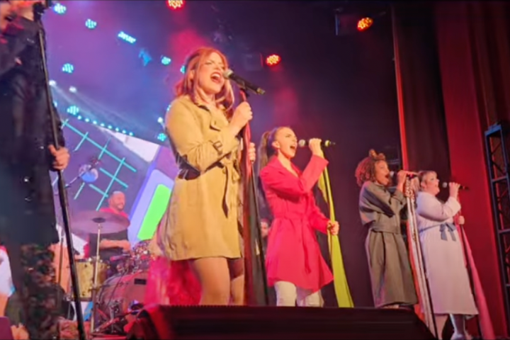 Singers on stage with colorful outfits, performing with microphones under bright lights.