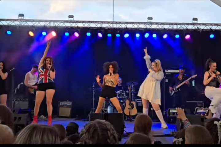 Five performers on stage singing and dancing with colorful lights.
