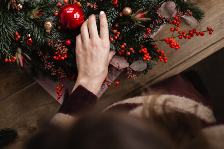 Person arranging a festive Christmas wreath with red berries and ornaments on a wooden table.