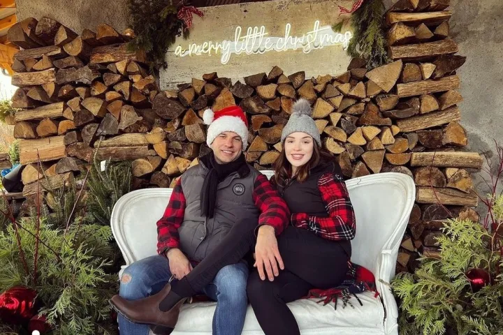Couple in festive hats sit on a couch with stacked wood and Christmas sign behind.