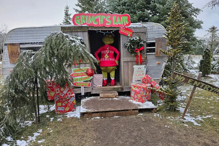 Grinch decor by a trailer with signs, gifts, and snow-covered trees.