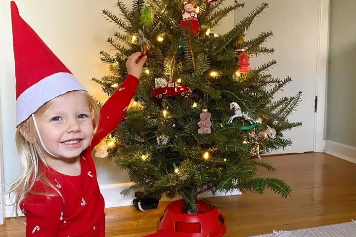 Child in red pajamas and hat decorates a small Christmas tree with ornaments and lights.